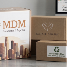 "A variety of custom-printed corrugated boxes for Los Angeles businesses by MDM Packaging, featuring a detailed LA skyline graphic, durable flexo print kraft boxes with 'Made in LA' text, and a sustainable packaging option with a high-recycled content label, all arranged on a desk.
